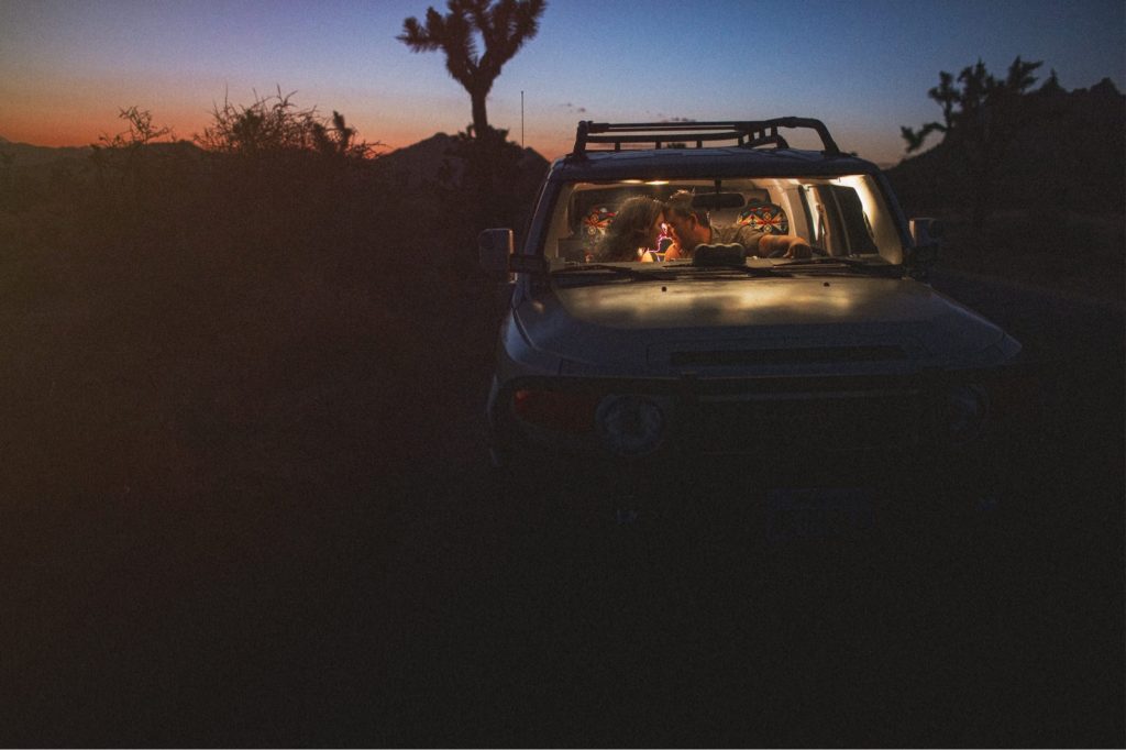 Sunset engagement session at Joshua Tree national park of couple in their Toyota FJ car kissing with light behind them