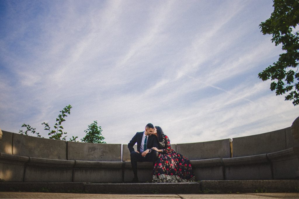 Boston Harbor walk Engagement session with creative couple sitting on bench and bride giving her groom a kiss