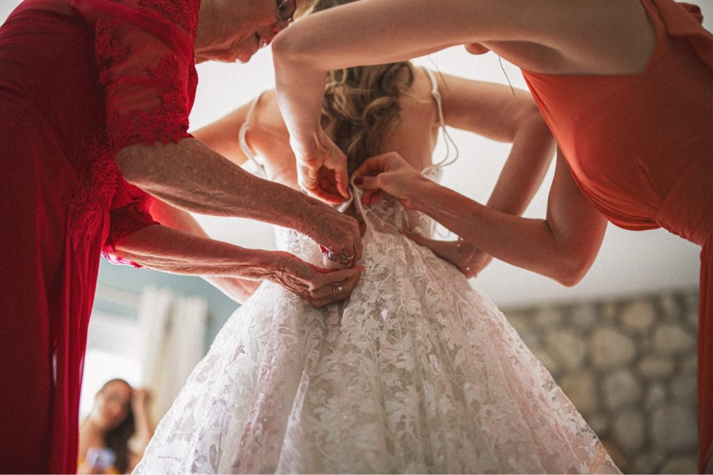 Photo of a bride getting ready and her mom and bridesmaid zipping up her dress from behind 