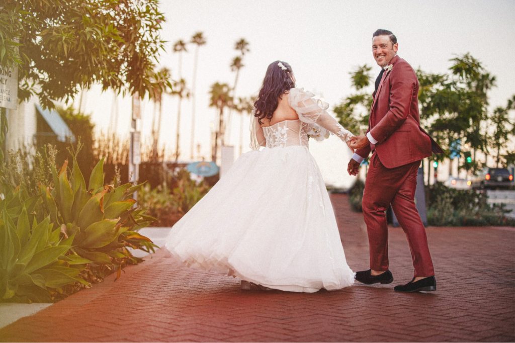 Hotel Californian Wedding with couple walking down state street in Santa Barbara before their reception with groom in his red suit looking back at camera smiling