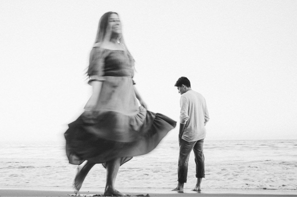 Black and white engagement session photo at butterfly beach in Montecito with bride twirling in foreground and groom stoic in the back ground 