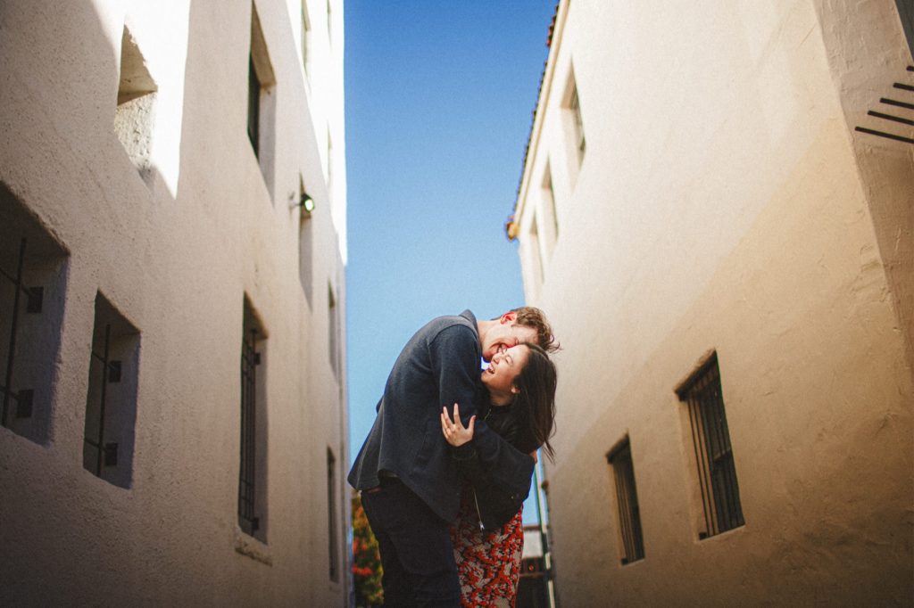 Creative photo of a down town Santa Barbara engagement session with couple holding each other laughing and being candid in between two buildings with the blue sky in the background