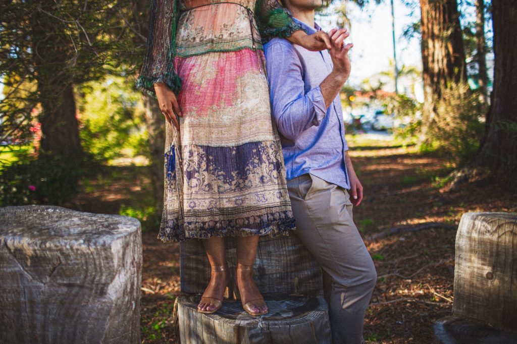 A creative engagement photo in Santa Barbara California taken at the Santa Barbara courthouse where the bride is standing on a wood stump holding the grooms hand while he leans up against her