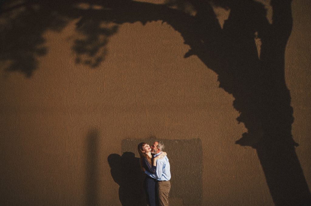 Creative portrait of a couple in Ventura on their engagement session up against a Wall with the shadow of a tree arching over them while they laugh and kiss
