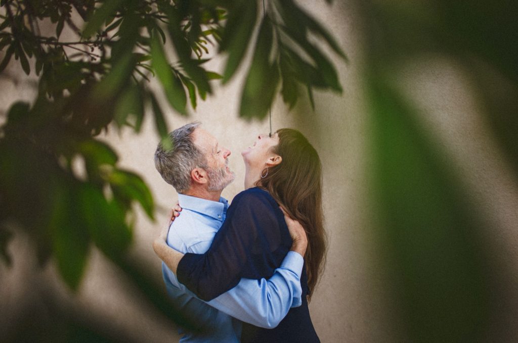 Couple hugging and laughing with tree leaves in the foreground wrapping around them while they have fun on their engagement session in Ventura