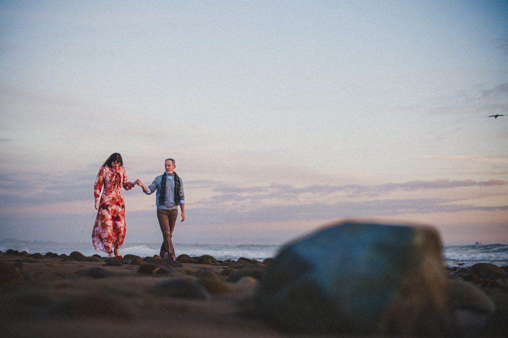 A couple walking in Ventura on their creative engagement session taken from the sand wild a couple walks across the beach holding hands at surfers point