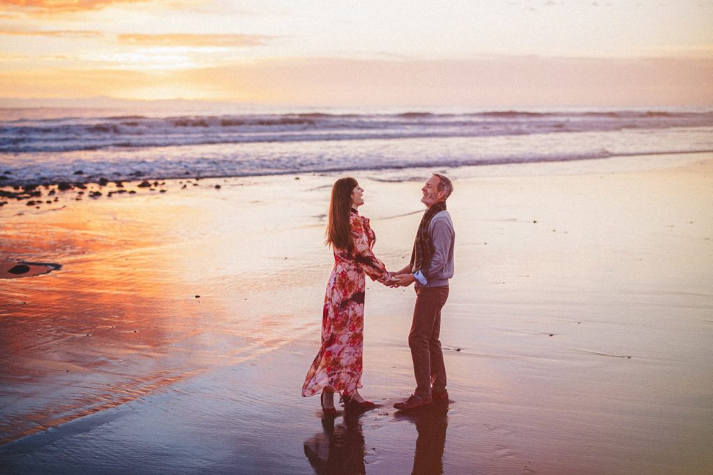 The couple holding hands at surfers point Beach on their engagement session in Ventura with a sunset behind them in the glassy water reflecting the beautiful colors
