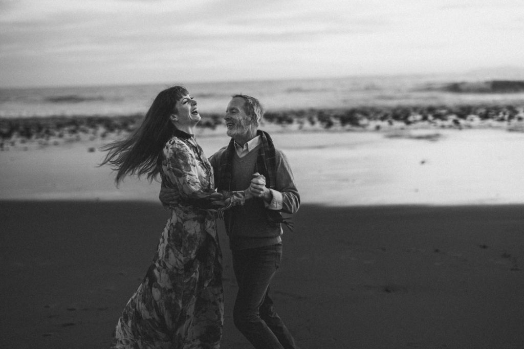 A classic and romantic black-and-white photo of a couple dancing on the beach and laughing as they have a candid moment